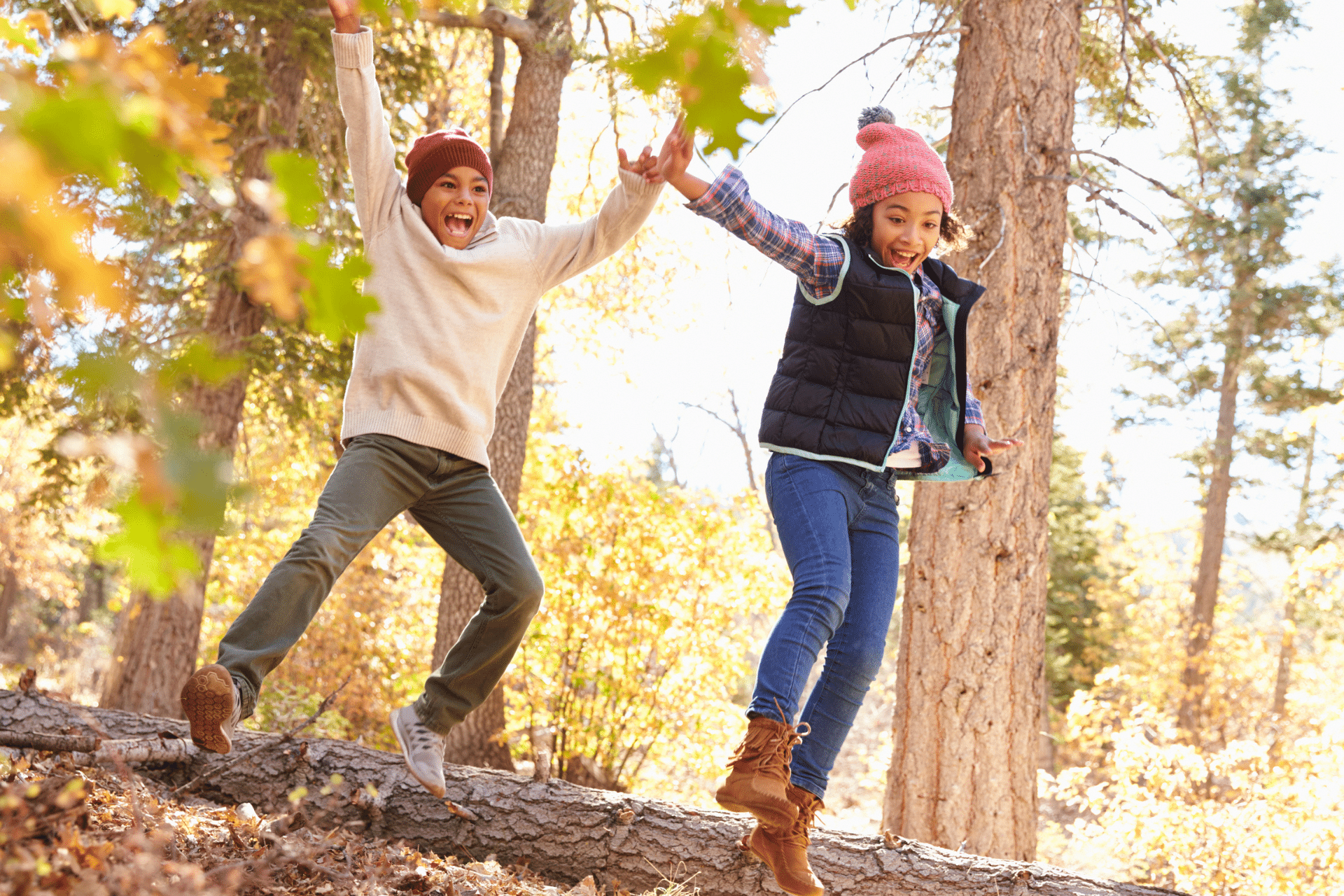 Two happy children wearing beanies jumping happily off a fallen tree log outdoors in autumn